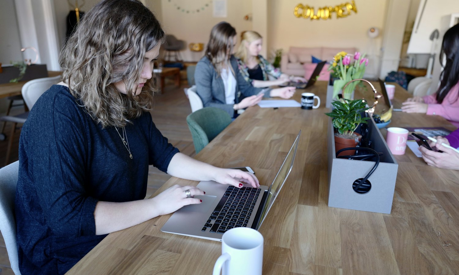 Femme qui travaille sur un ordinateur portable à une longue table de bois, des collègues derrière elle
