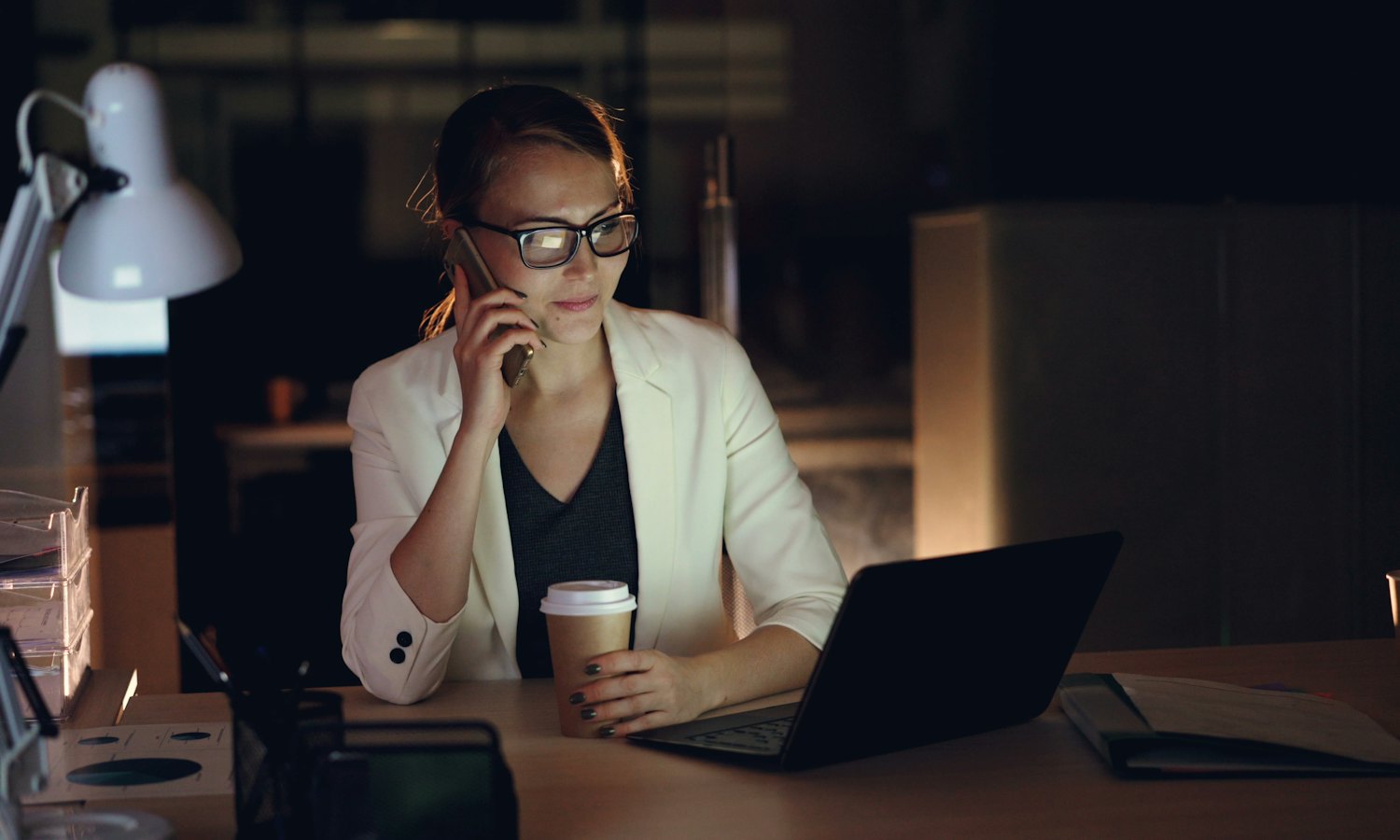 Femme au téléphone devant son ordinateur portable, le soir, une lampe allumée dans le bureau sombre