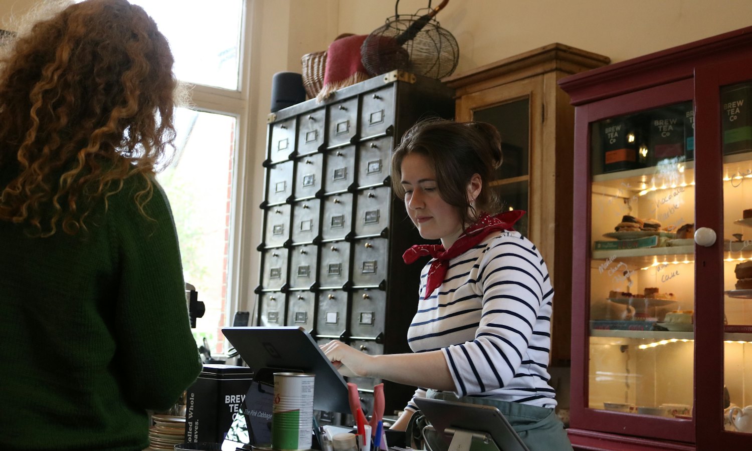 Commerçante à son comptoir, une tablette devant elle, devant une armoire à tiroirs et une vitrine